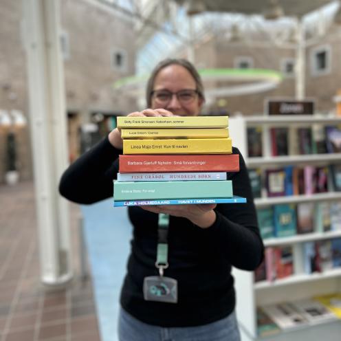 Tine står blandt fyldte bogreoler på Hovedbiblioteket. Hun holder syv af de omtalte bøger i denne artikel frem for sig i en stak med bogryggene frem. Der er fokus på bogryggene, mens både Tine og resten af baggrunden, som viser biblioteksrummet, fremstår let sløret. Man kan ane, at Tine smiler.
