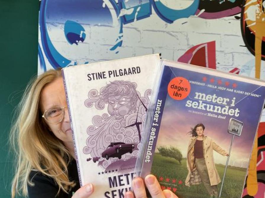 Lejla står op af en farverig væg. Hun holder "Meter i sekundet" som bog og film i hænderne foran sit ansigt..