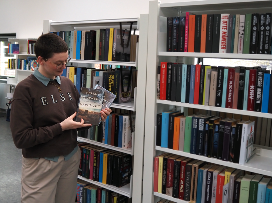 Anne Cecilie står med to af Helle Mogensens romaner i hænderne, som hun kigger ned på. Bag hende ses fyldte bogreoler og resten af biblioteksrummet.